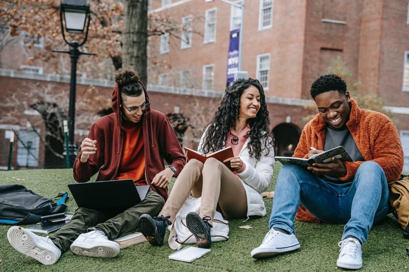 Three students sitting on grass field with laptop & books